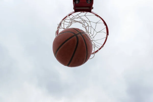 Orange ball inside red basketball hoop, basket against white sky. Outdoor basketball court.