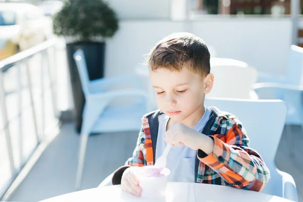 Menino bonito pré-escolar em camisa xadrez comer sorvete de morango em uma xícara sentada em um café em uma rua dia de verão — Fotografia de Stock