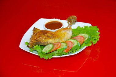 Kafeteryadaki fotoğrafta yemek, sebzeli garnitürlü kızarmış tavuk ve kırmızı arka planda sos var..