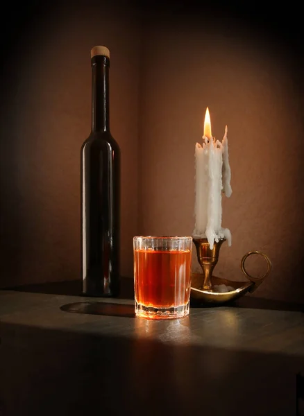 Still life with fruit liqueur in the vintage small glass, black bottle and candlestick with burning candle against a low key background. Selective and soft focus.