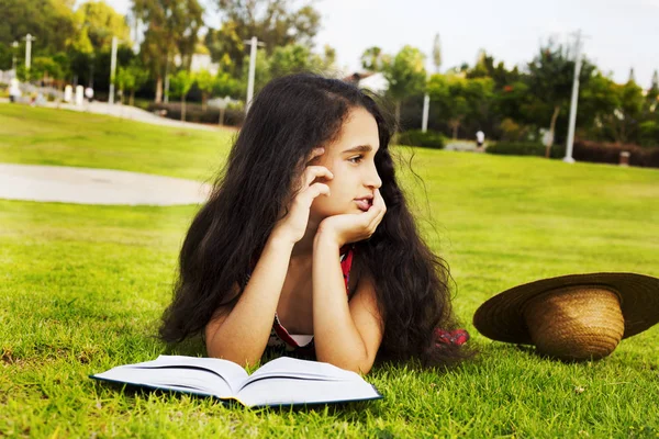 Das kleine afrikanische Mädchen, das mit Buch auf einem Gras im Park liegt und — Stockfoto