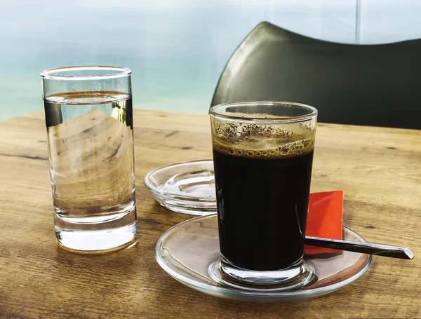 Sterke en donkere koffie in een glas geserveerd met een glas water op een houten tafel — Stockfoto