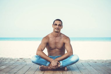 Kot pantolonlu genç kaslı adam sahilde dinleniyor ve poz veriyor.