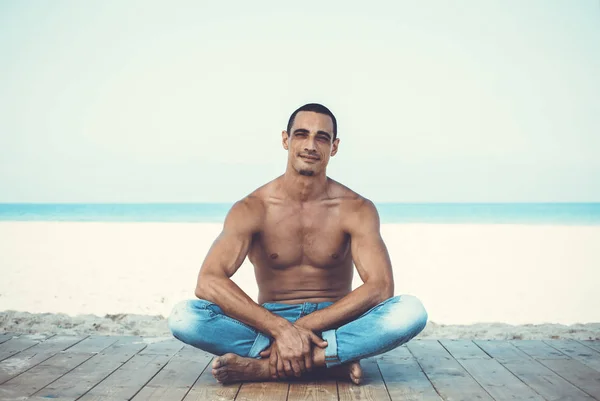 young muscular man in denim pants resting and posing on the beach