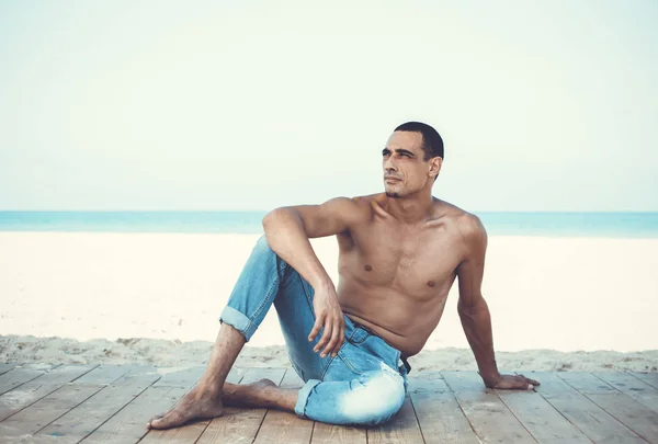 young muscular man in denim pants resting and posing on the beach