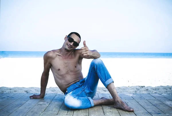 Joven musculoso en pantalones vaqueros descansando y posando en la playa — Foto de Stock