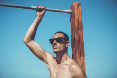 Genç kaslı adam sahilde dinleniyor ve poz veriyor. Güneş gözlüğü takıyor.