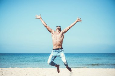 Genç kaslı adam sahilde dinleniyor ve poz veriyor. Havaya zıpla