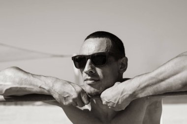 Genç kaslı adam sahilde dinleniyor ve poz veriyor. Güneş gözlüğü takıyor.