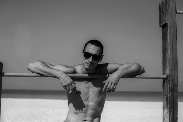 Joven musculoso descansando y posando en la playa. Usar gafas de sol — Foto de Stock