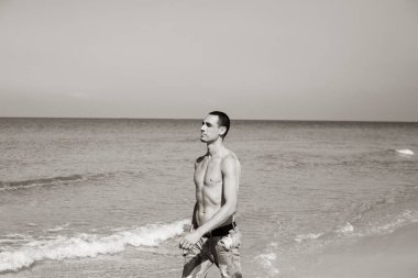 Genç kaslı adam sahilde dinleniyor ve poz veriyor. Genç bir adam deniz kenarında yürüyor.