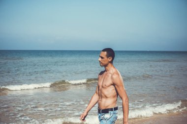 Genç kaslı adam sahilde dinleniyor ve poz veriyor. Genç bir adam deniz kenarında yürüyor.