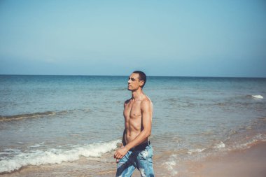 Genç kaslı adam sahilde dinleniyor ve poz veriyor. Genç bir adam deniz kenarında yürüyor.