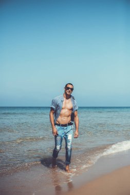 Genç kaslı adam sahilde dinleniyor ve poz veriyor. Genç bir adam deniz kenarında yürüyor.