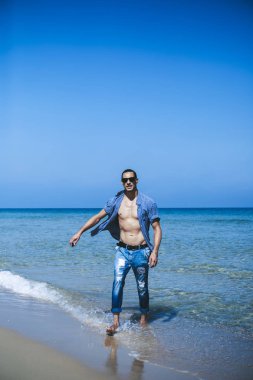 Genç kaslı adam sahilde dinleniyor ve poz veriyor. Genç bir adam deniz kenarında yürüyor.