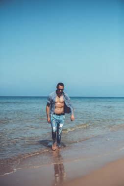 Genç kaslı adam sahilde dinleniyor ve poz veriyor. Genç bir adam deniz kenarında yürüyor.