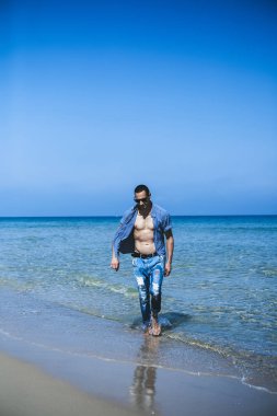 Genç kaslı adam sahilde dinleniyor ve poz veriyor. Genç bir adam deniz kenarında yürüyor.