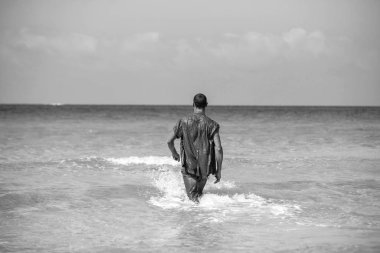 Genç kaslı adam sahilde dinleniyor ve poz veriyor. Genç bir adam deniz kenarında yürüyor.