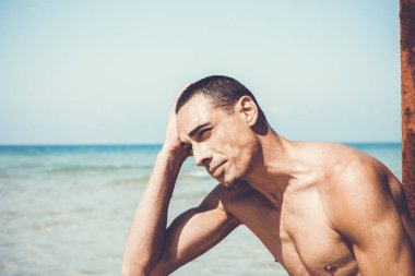 Genç kaslı adam sahilde dinleniyor ve poz veriyor.