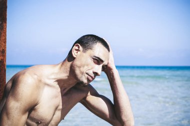 Genç kaslı adam sahilde dinleniyor ve poz veriyor.