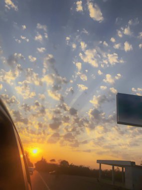 Gün batımında bulutlu bulutlu gökyüzü sıcak gün batımı ışığıyla aydınlatılıyor. Gün batımında köy yolu