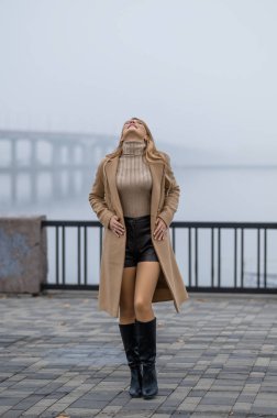 Uzun saçlı, çekici bir sarışın sabahları sonbahar gezintisi boyunca yürür. Sis. Palto ve çizmeli sarışın. Bir sonbahar fotoğraf çekimi için poz vermek