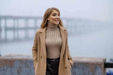 Uzun saçlı, çekici bir sarışın sabahları sonbahar gezintisi boyunca yürür. Sis. Palto ve çizmeli sarışın. Bir sonbahar fotoğraf çekimi için poz vermek