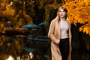 Sonbaharda, bej ceketli güzel bir sarışın parkta yürür. Sarı yapraklar, sonbahar, doğa. Ormanda sonbahar fotoğraf çekimi