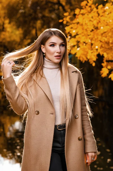 公園の秋にはベージュのコートを着た美しいブロンドが歩いています 黄色の葉 森の秋の撮影 — ストック写真