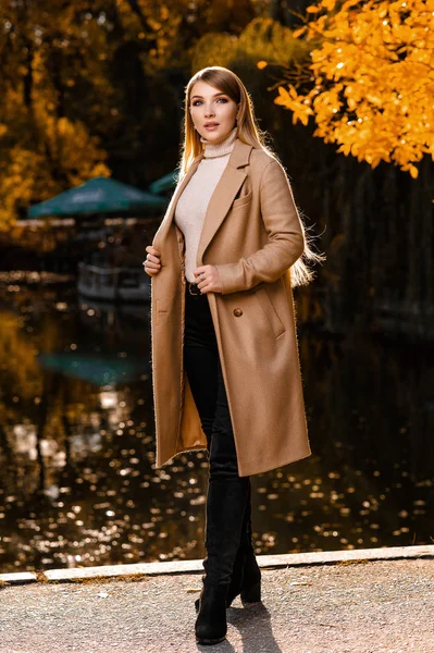 Sonbaharda, bej ceketli güzel bir sarışın parkta yürür. Sarı yapraklar, sonbahar, doğa. Ormanda sonbahar fotoğraf çekimi