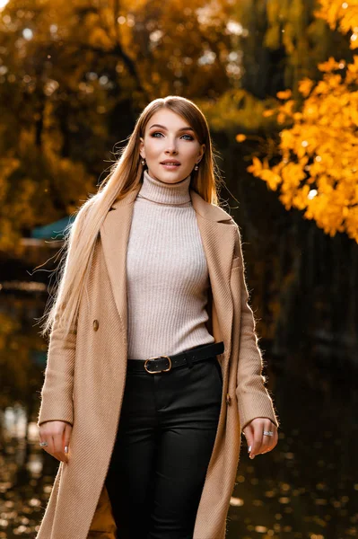 Sonbaharda, bej ceketli güzel bir sarışın parkta yürür. Sarı yapraklar, sonbahar, doğa. Ormanda sonbahar fotoğraf çekimi