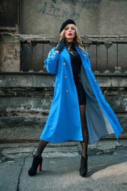 Mavi pelerinli bir sarışın cadde boyunca binaların arka planında yürüyor.