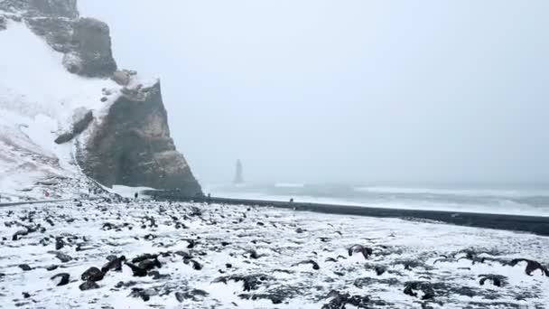 Vista aérea de uma praia vulcânica preta e montanhas à beira-mar na Islândia. Baixo alcance sobre as rochas. A neve cai. Multidão de turistas — Vídeo de Stock