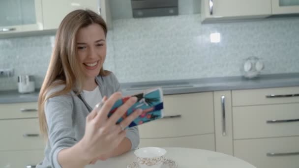 Chica atractiva con el pelo rojo bebe café de la mañana en la cocina del hogar. Hace una selfie en un smartphone para redes sociales. Mujer bonita usando el teléfono para foto selfie en la cocina moderna — Vídeos de Stock