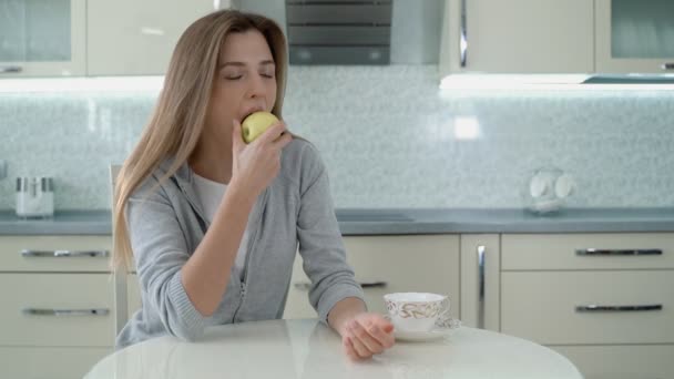 Una joven se sienta en casa en una cocina luminosa y se come una manzana . — Vídeo de stock