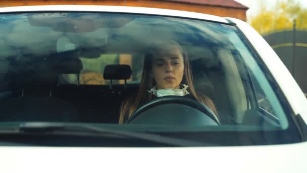 Ένα νεαρό κορίτσι με γάντια βάζει μια μάσκα του ιού στο πρόσωπό της ενώ κάθεται σε ένα αυτοκίνητο, με θέα μέσα από ένα παρμπρίζ — Αρχείο Βίντεο