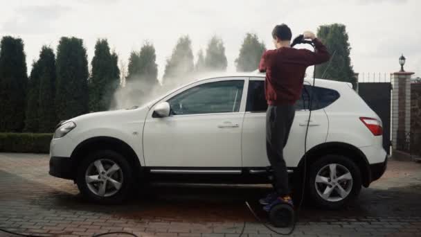 Dospělý muž mytí svého bílého auta v myčce aut pomocí tlakové myčky venku na večer. — Stock video