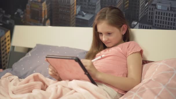 Gyönyörű lány fekszik az ágyban, és lefekvés előtt, és rajzol egy tablettát ceruzával — Stock videók