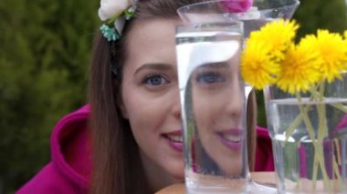 Bir kız kameraya bakar, su dolu bir bardak suyun içinden, çerçevede bahar çiçekleri karahindiba, kız yüzünü suyla dolu bir bardağın arkasına saklar..