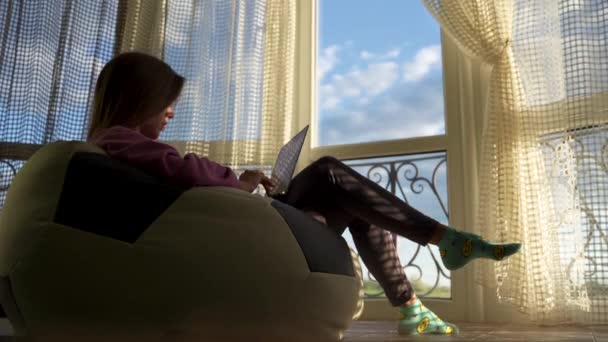 Самоизоляция Covid-19 и работа из дома. Женщина с ноутбуком сидит на мягком диване возле большого окна — стоковое видео
