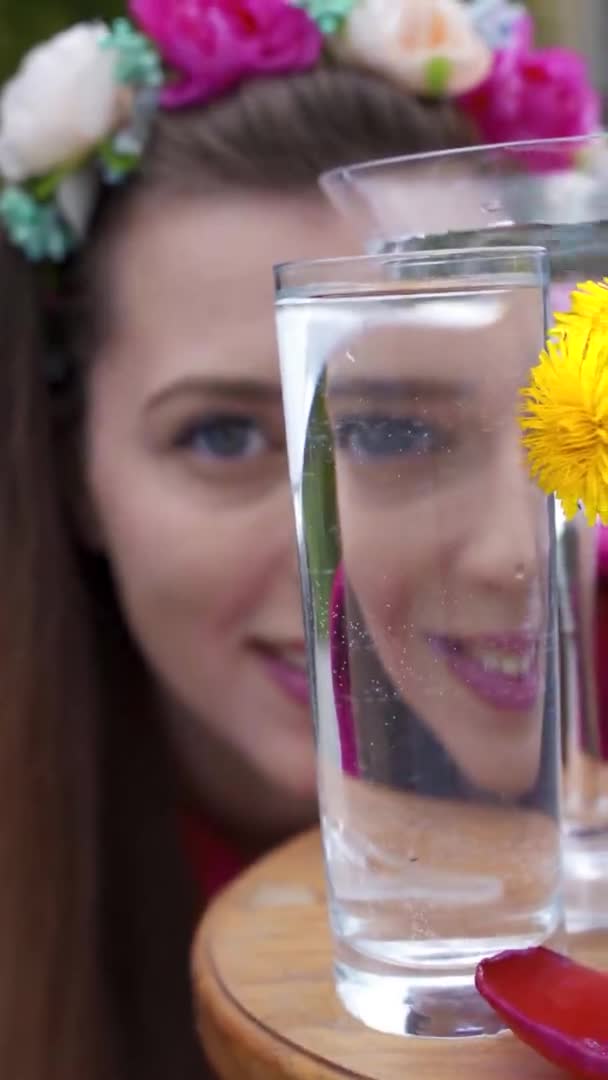 Een meisje kijkt in de camera, door een glas water dat gevuld is met water, in het frame lentebloemen paardebloemen, meisje verbergt haar gezicht achter een glas met water. — Stockvideo