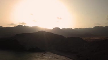 Hava aracı gün batımında turuncu gökyüzüne karşı deniz kenarındaki kayalık dağlarda hareket ediyor.