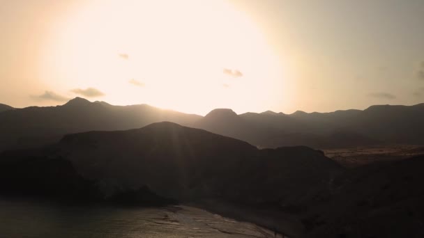 Antenne: Drohne bewegt sich bei Sonnenuntergang über silhouette felsige Berge am Meer gegen orangefarbenen Himmel — Stockvideo