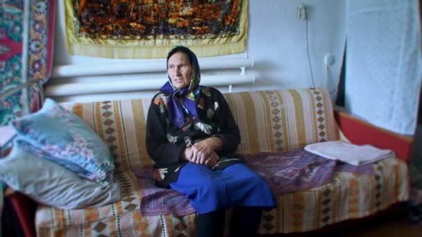 Una anciana en un pañuelo brillante sentado en la habitación en el sofá — Vídeos de Stock