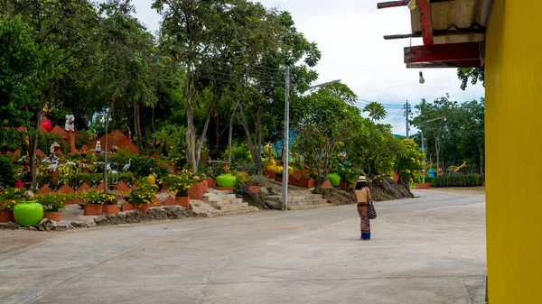 Woman in sarong and fedora walking down road in wat with garden to her left