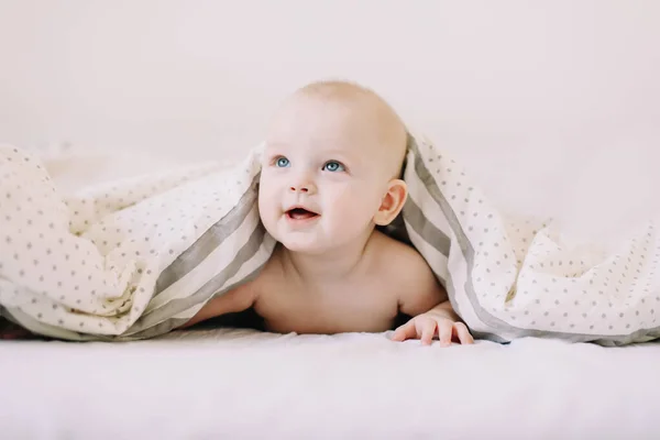 Sevimli bebek beyaz yatak odasında emeklemeyi öğreniyor. Yatakta battaniyenin altında yatan sevimli komik bebek. Karnını doyuran küçük çocuk. — Stok fotoğraf