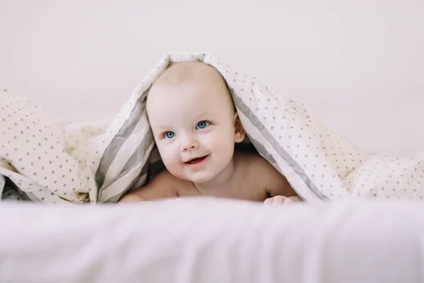 Adorable bebé aprendiendo a gatear en el dormitorio blanco. Lindo bebé divertido acostado debajo de la manta en la cama. Pequeño niño haciendo tiempo de barriga — Foto de Stock