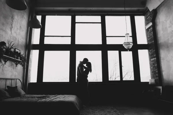 Silhueta de jovem casal de pé e abraçando contra fundo janela dentro de casa. retrato de jovem belo casal . — Fotografia de Stock