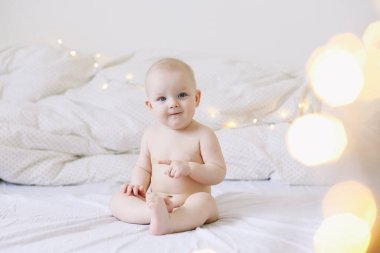 Sevimli gülümseyen bebek yatağa oturur. Mutlu çıplak bebek. Bir yaşın altındaki çocuklar. Bebek kız oynuyor, gülümsüyor, yatakta poz veriyor. 