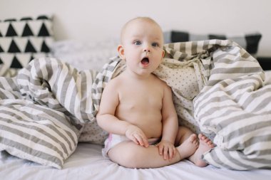 Sevimli gülümseyen bebek yatağa oturur. Mutlu çıplak bebek. Bir yaşın altındaki çocuklar. Bebek kız oynuyor, gülümsüyor, yatakta poz veriyor. 
